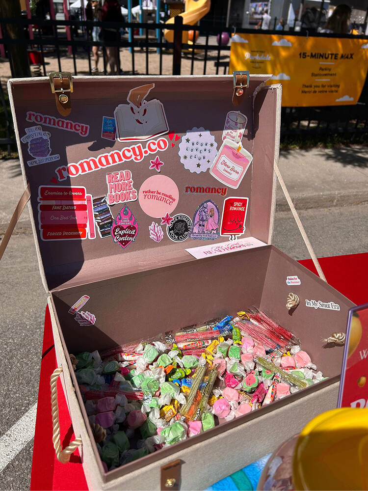 An open fabric trunk filled with colorful wrapped candies on an outdoor table. The inside lid is decorated with stickers featuring slogans like "READ MORE BOOKS," "Smell the Smut," "ENEMIES TO LOVERS," and "ROMANCY.CO".