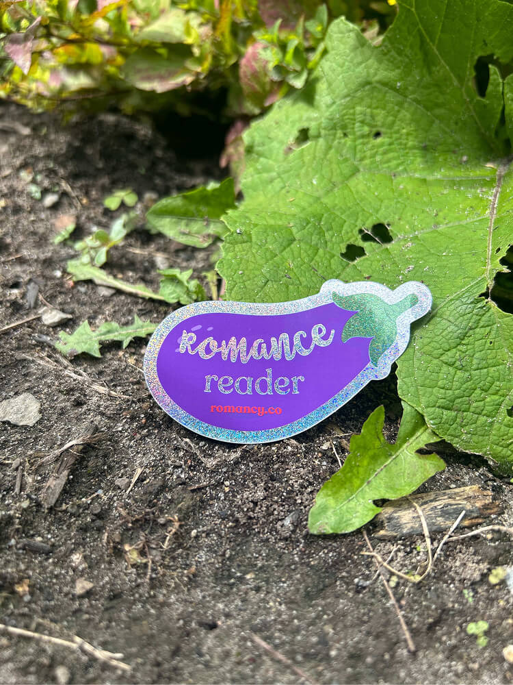 A purple eggplant-shaped glittery sticker resting on soil next to a large green leaf, reading "Romance reader" and "romancy.co".