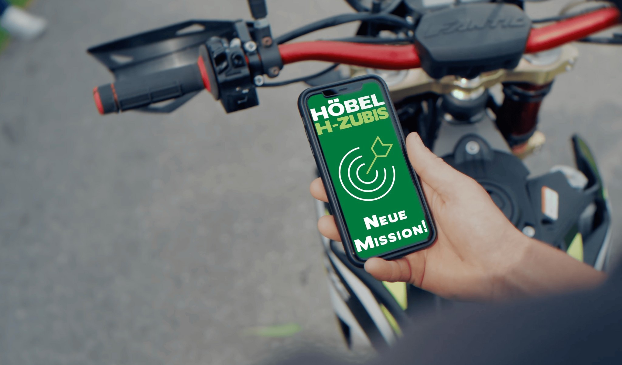 Hand hält Smartphone mit Höbel-Azubi-Kampagne „Neue Mission“