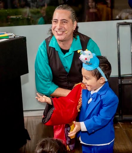 JamesZini smiling as a young magician in a blue suit helps perform a magic trick