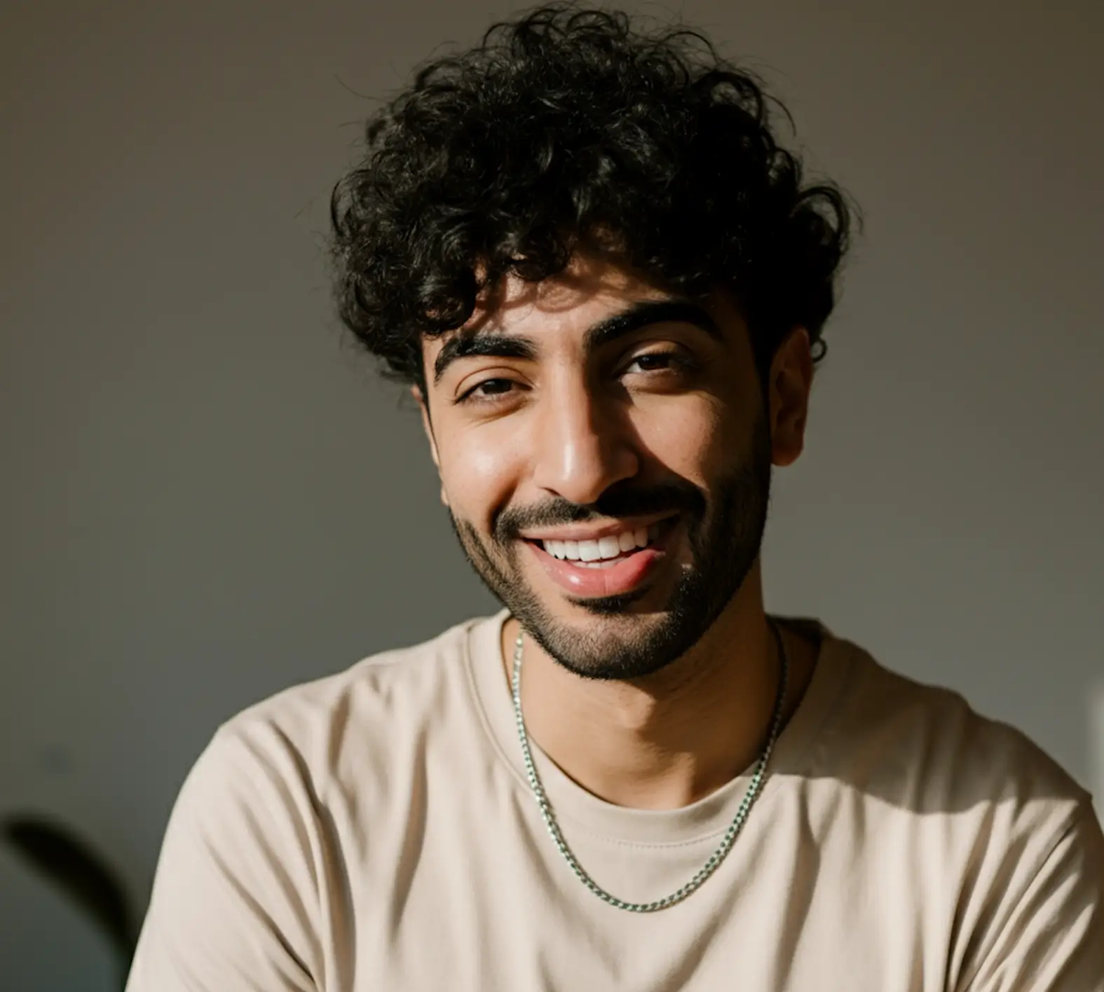 Smiling man with curly dark hair, beard, and mustache, wearing a beige shirt and silver chain necklace against a plain background.