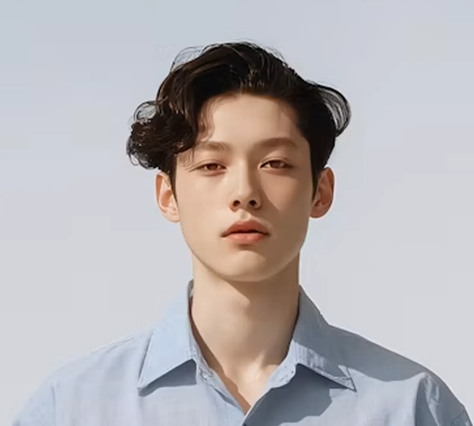 Close-up of a young man with wavy dark hair wearing a light blue button-up shirt, looking directly at the camera against a plain light background.