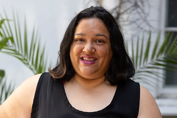 Smiling woman with shoulder-length dark hair wearing a black sleeveless top in an outdoor setting with green palm leaves in the background.