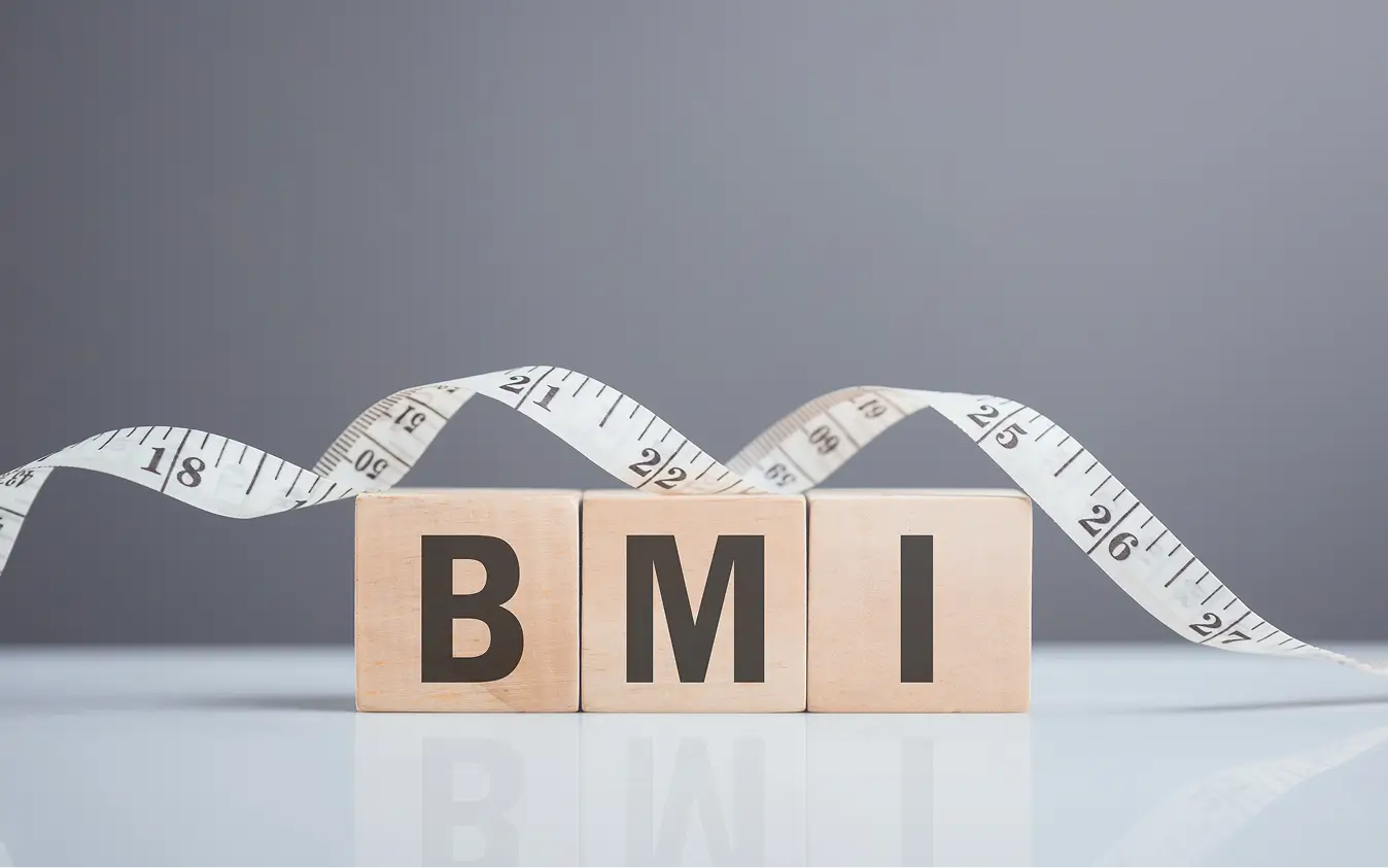 Wooden blocks spelling out BMI with a measuring tape draped over them on a reflective surface.