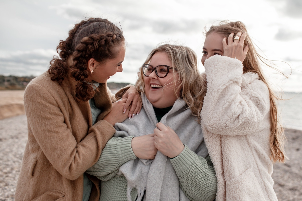 Three women dressed in winter coats laughing and embracing on a beach with a cloudy sky.