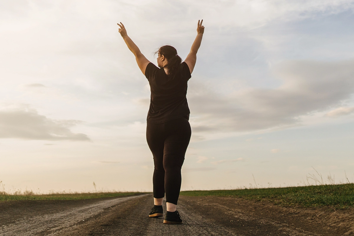 Woman standing on a dirt road with arms raised making peace signs against a cloudy sky.