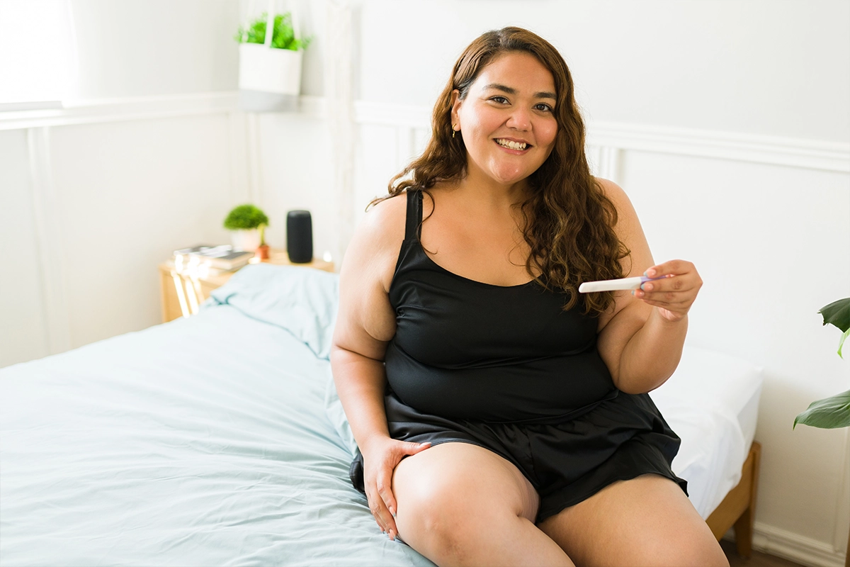 Smiling woman sitting on a bed holding a pregnancy test in a bright bedroom.