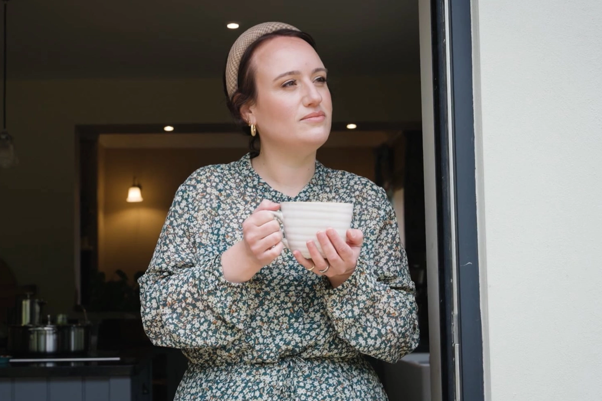 Woman with a headband holding a white mug and looking thoughtfully outside from a doorway.