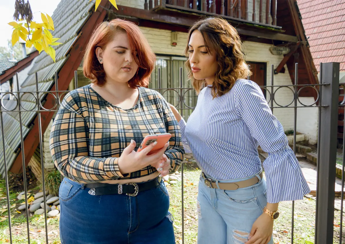 Two women standing outdoors by a metal fence, looking at a smartphone held by the woman with red hair wearing a plaid shirt and jeans.