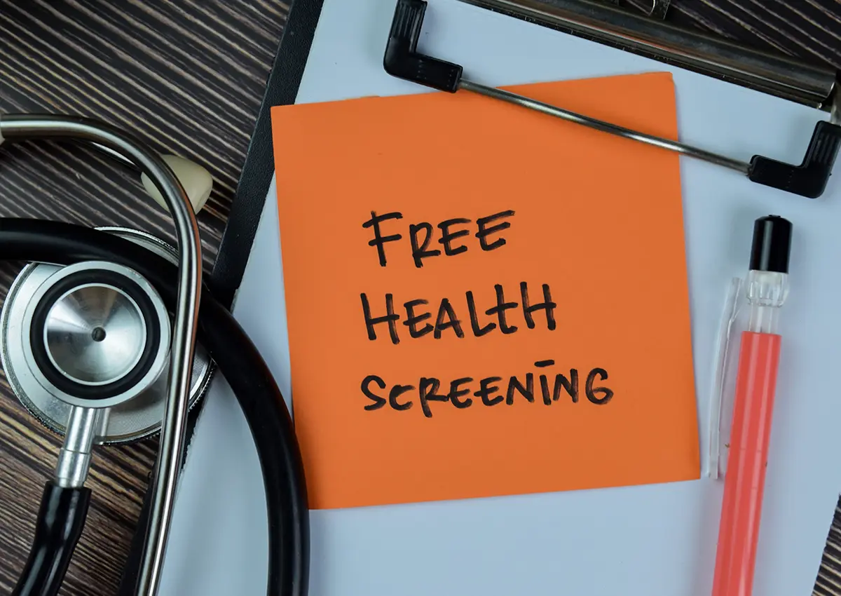 Orange sticky note on a clipboard with 'FREE HEALTH SCREENING' written on it, next to a stethoscope and a pink pen.
