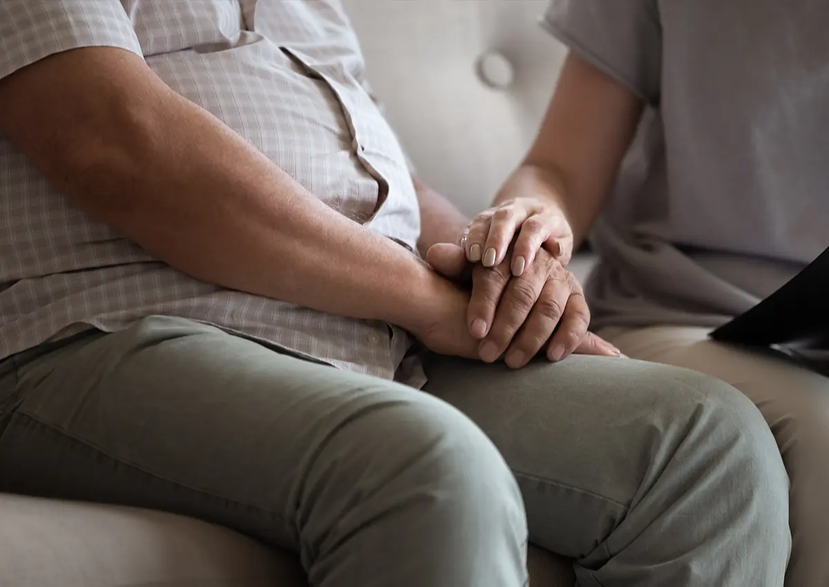 Close-up of two people sitting side by side, one holding the other's hand gently on their lap.