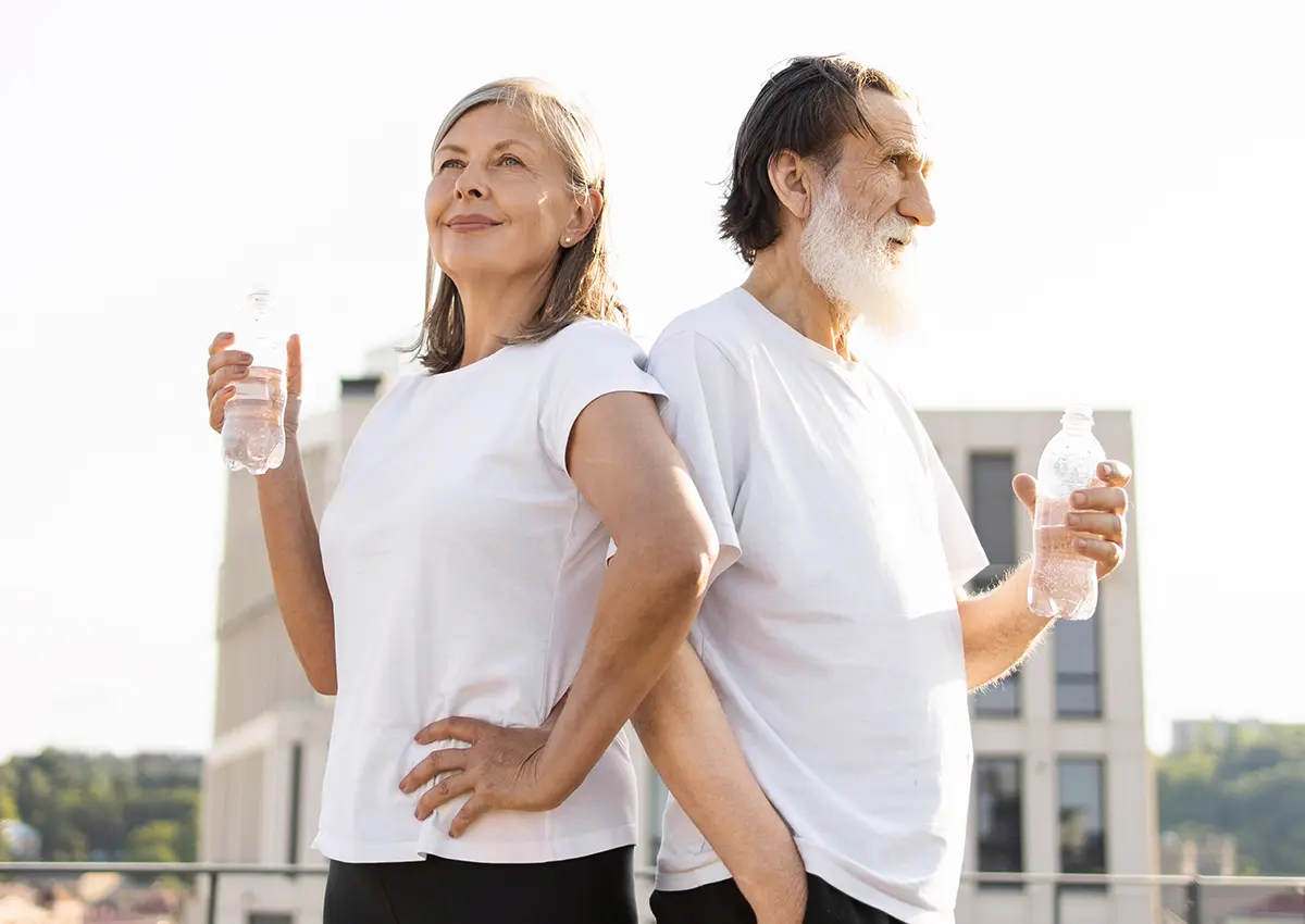 Older man and woman dressed in white t-shirts standing back to back, each holding a water bottle outdoors.