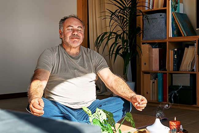 Middle-aged man with closed eyes meditating cross-legged in a sunlit room with plants and shelves.