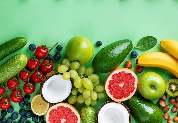 An assortment of fresh fruits and vegetables including bananas, green apples, avocados, tomatoes, grapes, blueberries, kiwis, lemons, coconuts, zucchinis, and hazelnuts on a green background.