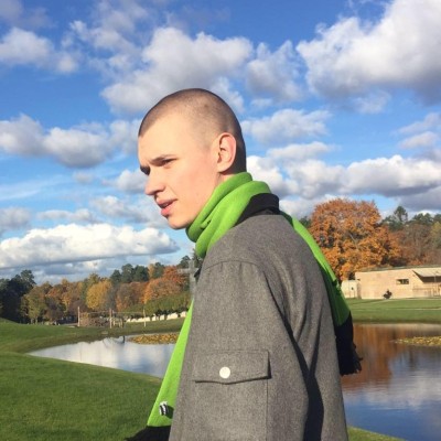 Young man with a shaved head wearing a grey coat and green scarf standing near a pond with trees and a partly cloudy sky in the background.
