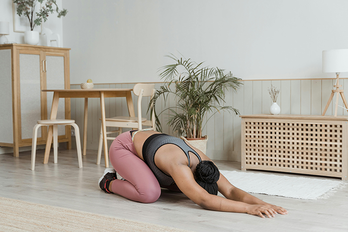 Woman in pink leggings and dark tank top performing a child's pose yoga stretch on a light wooden floor in a modern room.