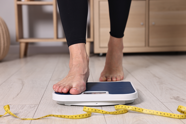 Person standing barefoot on a blue and white bathroom scale with a yellow measuring tape on the floor.