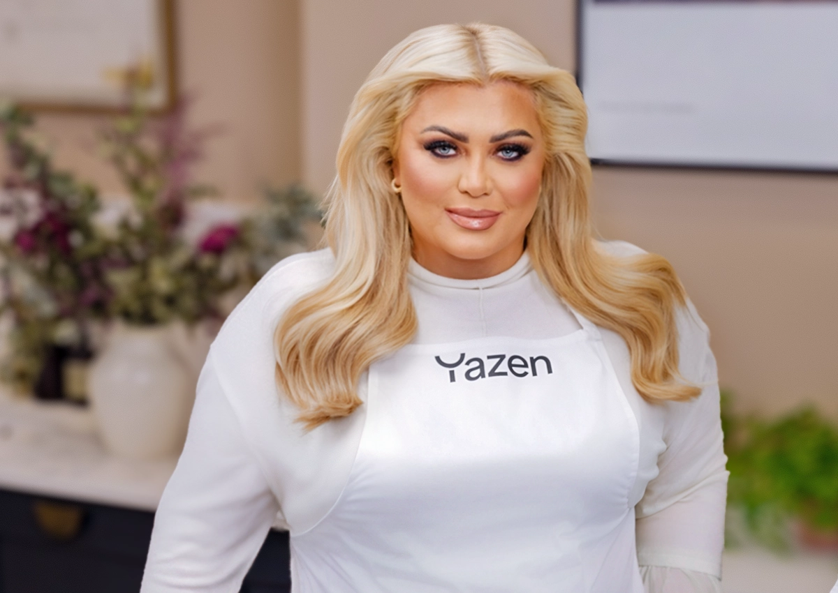 Woman with long blonde hair wearing a white shirt and apron with Yazen logo, indoors with blurred background.