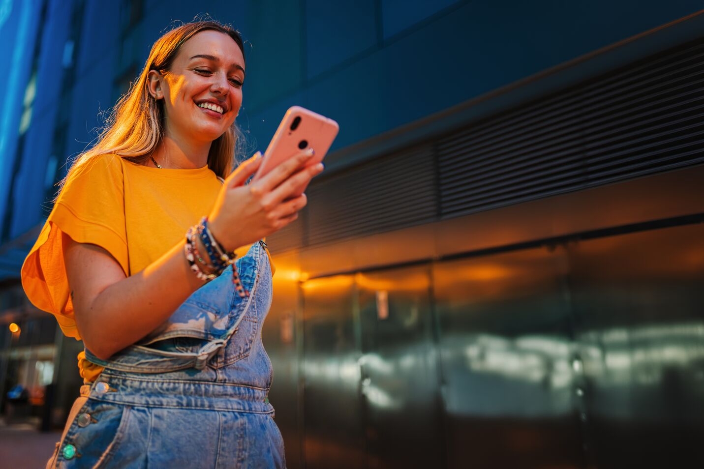 Smiling young woman wearing an orange shirt and denim overalls, standing outdoors at dusk while looking at her smartphone.