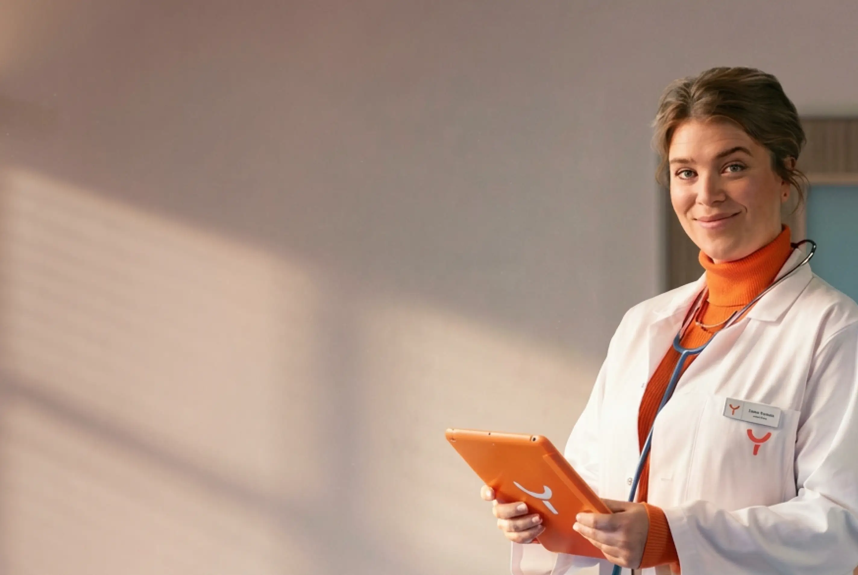 Smiling female doctor in a white coat with a stethoscope around her neck holding an orange tablet device.