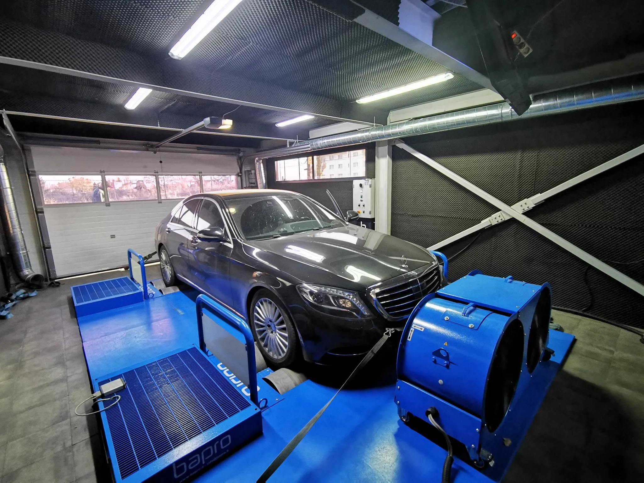 Black luxury sedan secured on a blue automotive dynamometer testing platform inside a garage.