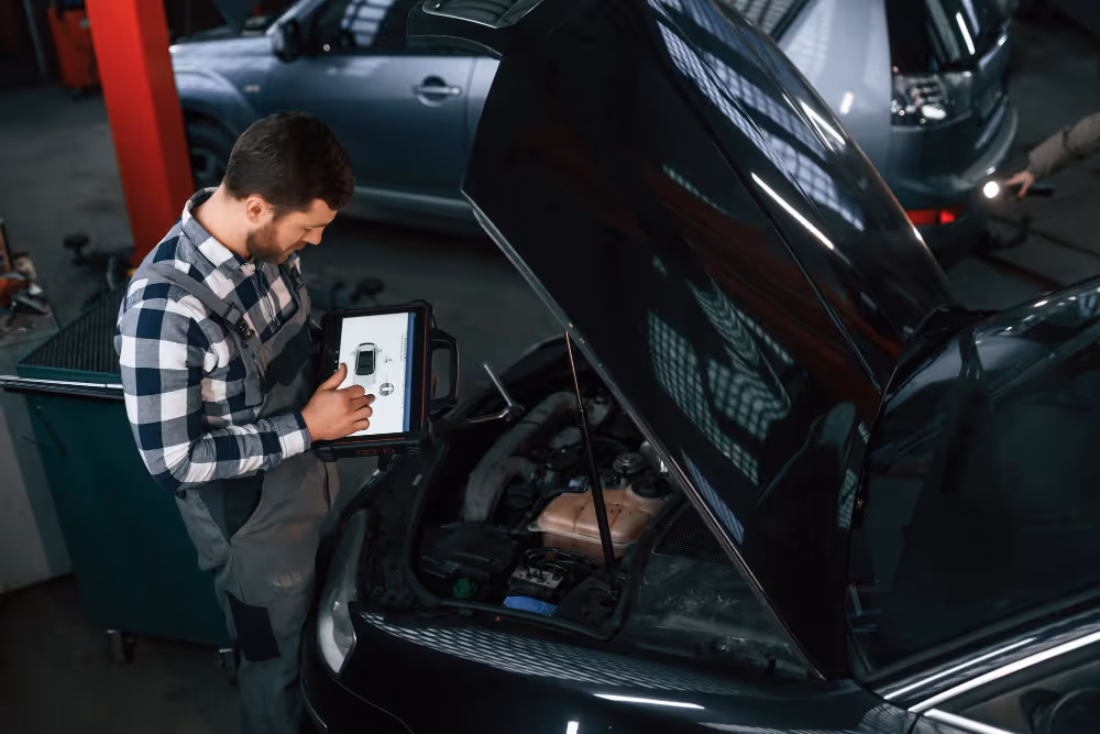 Mechanic using a diagnostic tablet to check a car engine with the hood open in a garage.