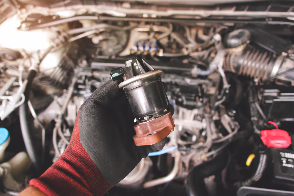 Gloved hand holding a car engine part with engine components blurred in the background.