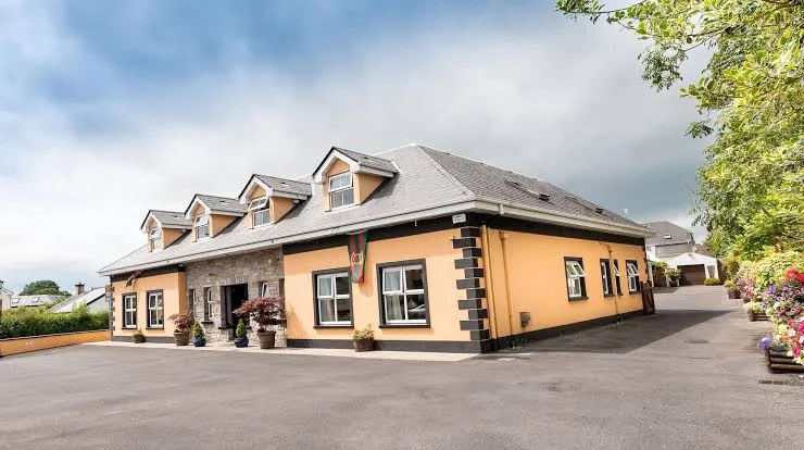 Single-story building with a gray roof and peach-colored walls featuring four dormer windows and decorative black corner accents, surrounded by paved driveway and landscaped greenery with flowers.
