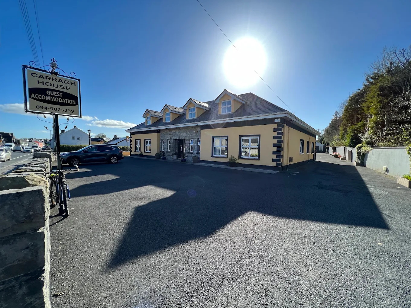 Carragh House guest accommodation building under bright sunlight with a clear blue sky, cars parked nearby, a bicycle near a stone wall, and trees on the right side.