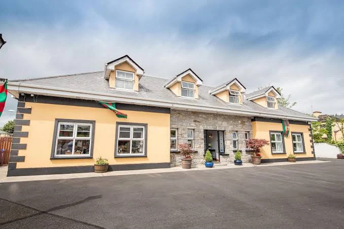 Wide front view of a single-story building with a gray roof featuring four dormer windows and beige walls accented with brown trim, with potted plants near the entrance and multiple windows reflecting the surroundings under a cloudy sky.
