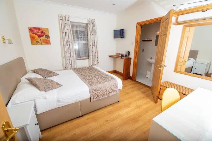 Clean, modern hotel room featuring a double bed with white linens and beige floral runners and pillows, a window with floral curtains, a mounted flat-screen TV, a small desk area with a kettle, and an open door leading to a bathroom with a sink and toilet. The room has light wood flooring, white walls, and a large mirror above a wooden bench beside a white desk and yellow chair.