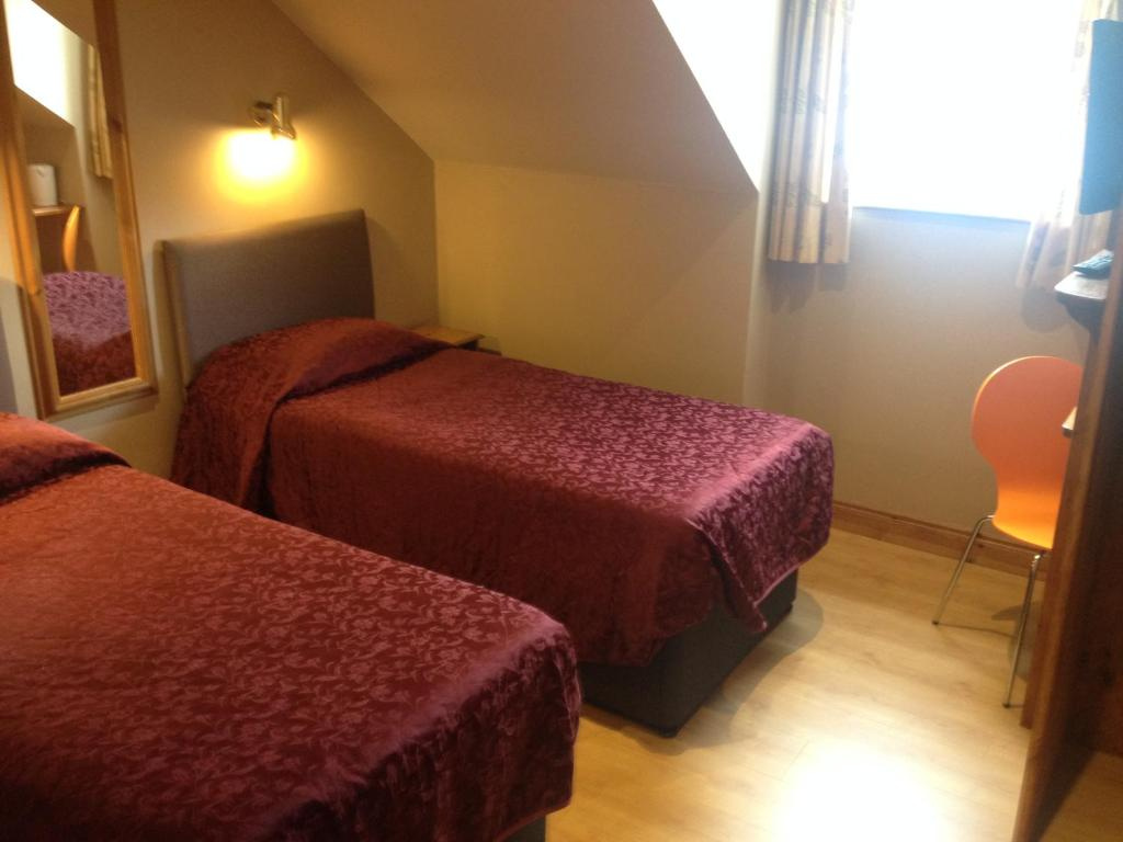 Small bedroom with two single beds covered in matching dark red patterned bedspreads, a wooden floor, wall-mounted light above one bed, a mirror on the wall, and a window with curtains; an orange chair is positioned near the window beside a mounted TV.