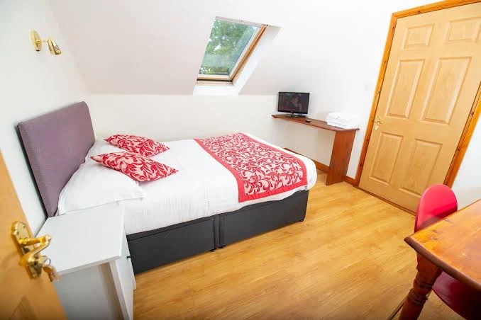 Small, bright bedroom with a double bed dressed in white linens, red and white patterned pillows and bedspread, a slanted skylight window above the bed, a wooden door, a white nightstand, a mounted wooden shelf holding a small TV and folded towels, and a corner of a wooden desk with a red chair.