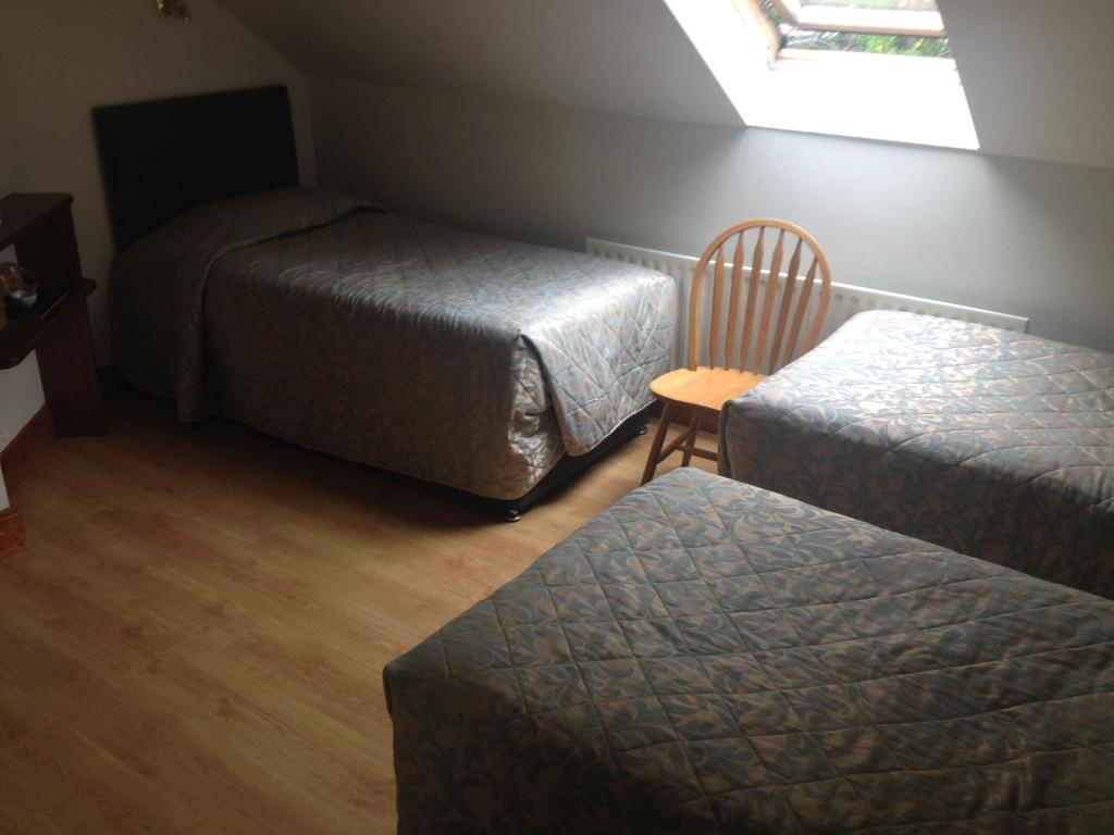 Bedroom with three single beds covered in patterned gray bedspreads arranged in an L shape near a slanted ceiling with a skylight window, and a wooden chair between two beds on wooden flooring.