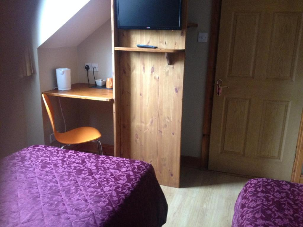 Small room with two beds covered in purple floral bedspreads, a wooden door, a built-in wooden panel with a mounted TV and remote, and a corner desk with an orange chair and electric kettle.