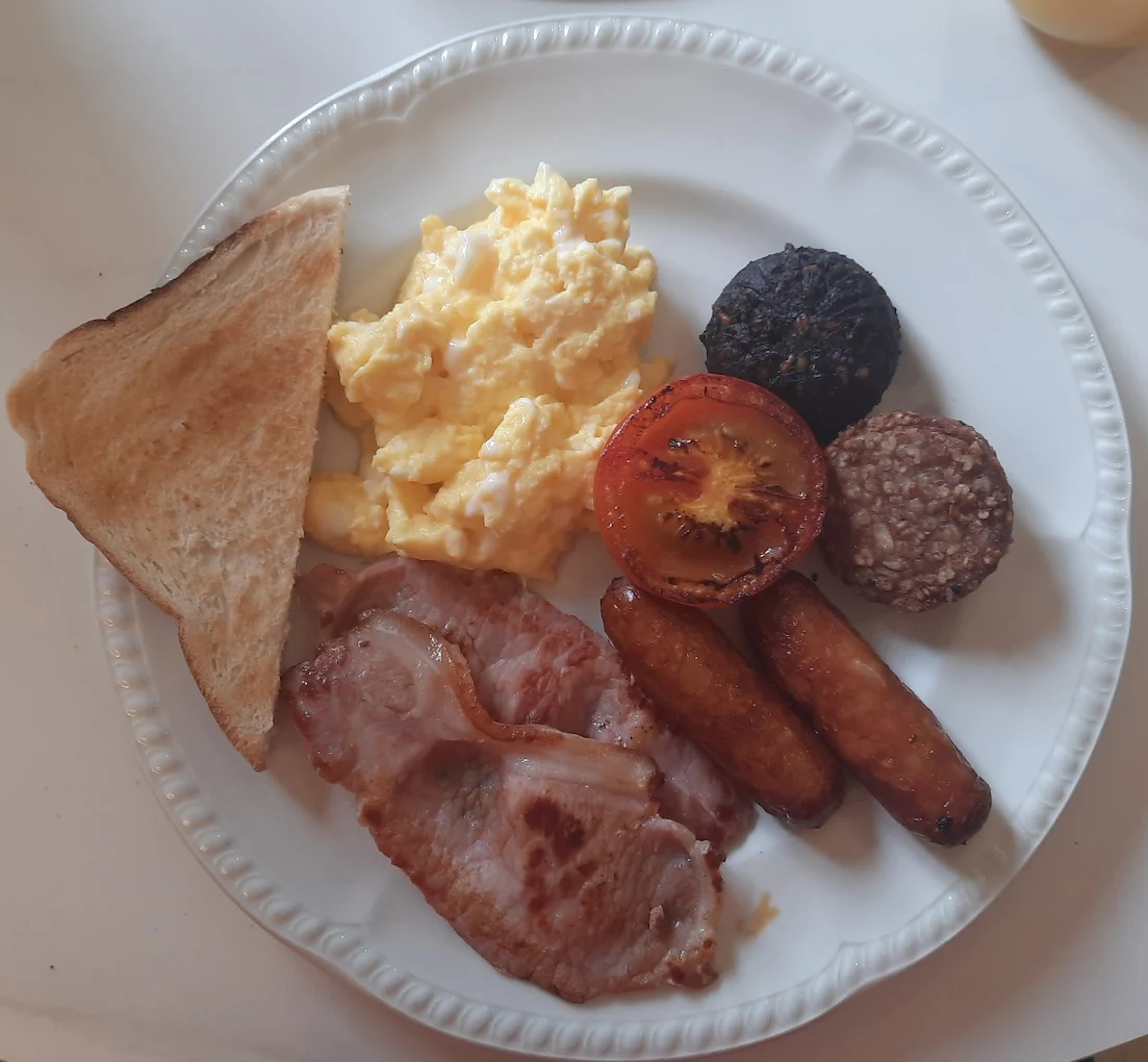 A plate of cooked breakfast featuring scrambled eggs, two slices of bacon, grilled tomato half, two breakfast sausages, two types of round black and white pudding, and a triangular slice of toasted bread.