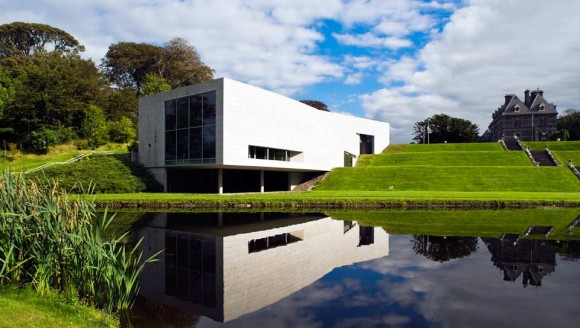Modern white building with large glass windows on a grassy slope, reflected in a calm pond with surrounding greenery and a partly cloudy sky.