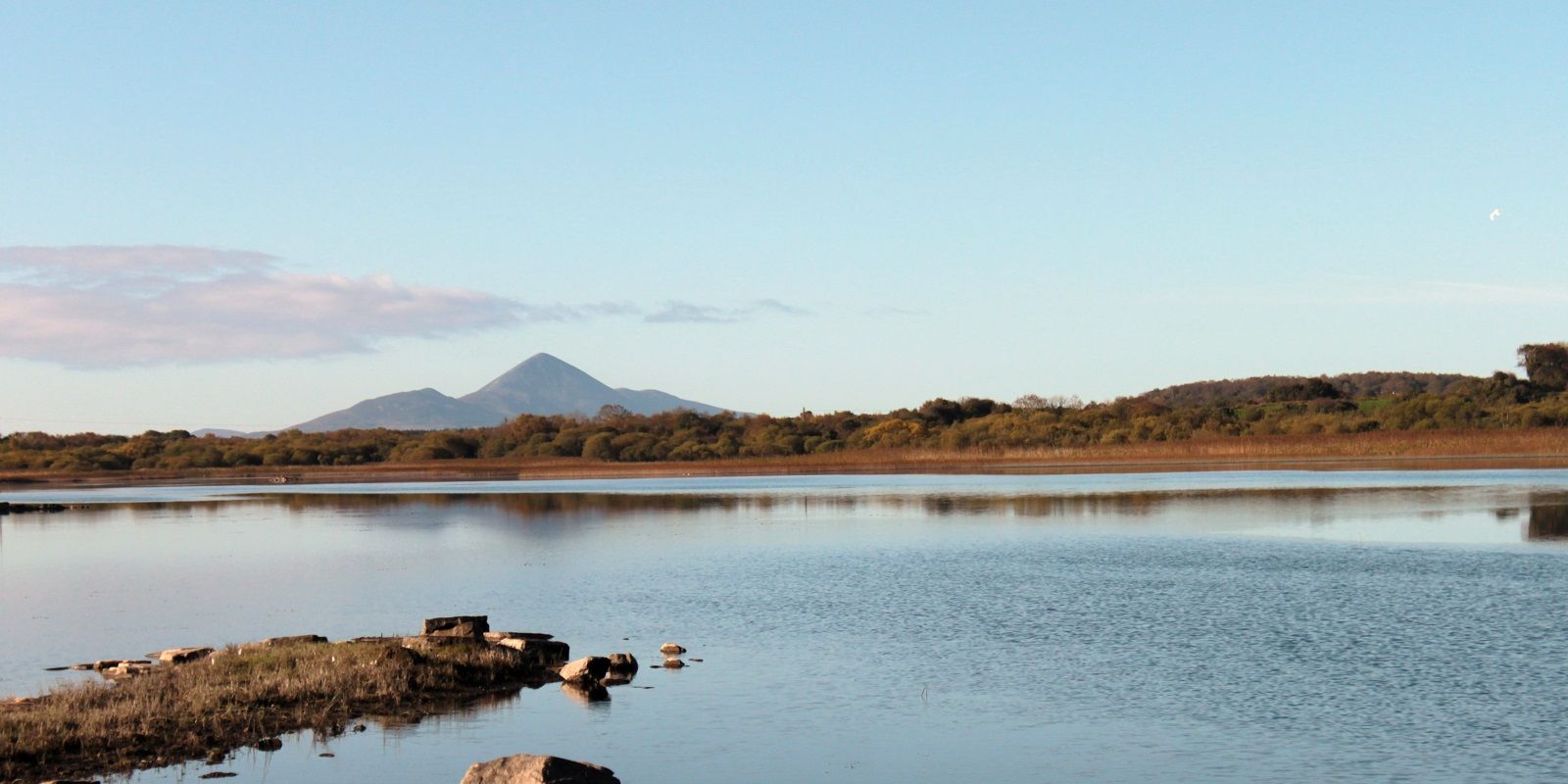 Calm body of water with a small rocky and grassy outcrop in the foreground, surrounded by a tree-covered shoreline and distant mountains under a clear blue sky.