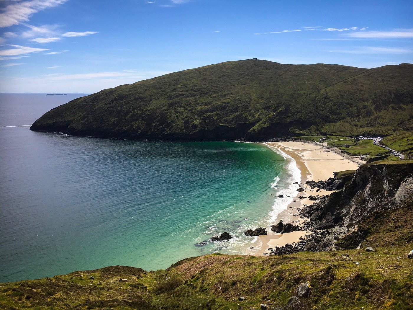 A scenic cove with turquoise water gently meeting a sandy beach surrounded by green, rocky hills under a blue sky with wispy clouds.