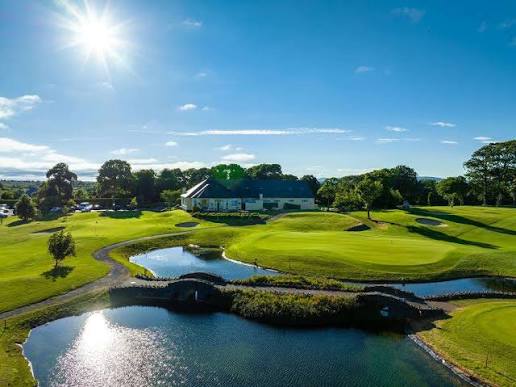 Sunny golf course with green fairways, a water hazard with a small stone bridge, and a clubhouse building surrounded by trees under a clear blue sky.