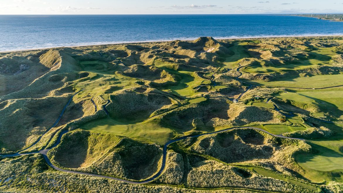 Aerial view of a coastal golf course with rolling sandy dunes, green fairways, and a winding path, bordered by the ocean under a partly cloudy sky.
