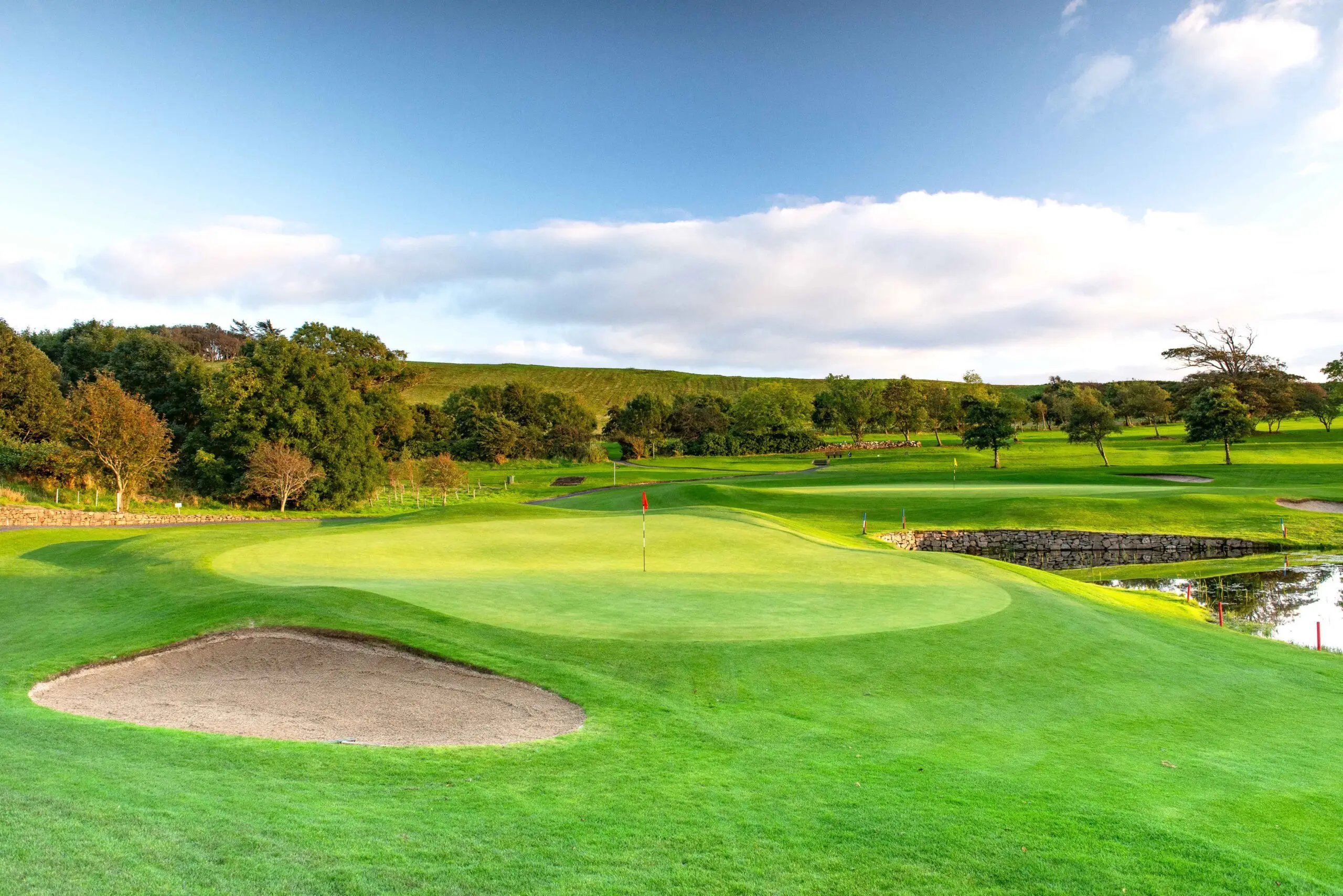 A lush green golf course with two putting greens marked by red and yellow flags, a sand bunker in the foreground, a stone wall near a water hazard reflecting nearby trees, and a clear blue sky with some clouds above.