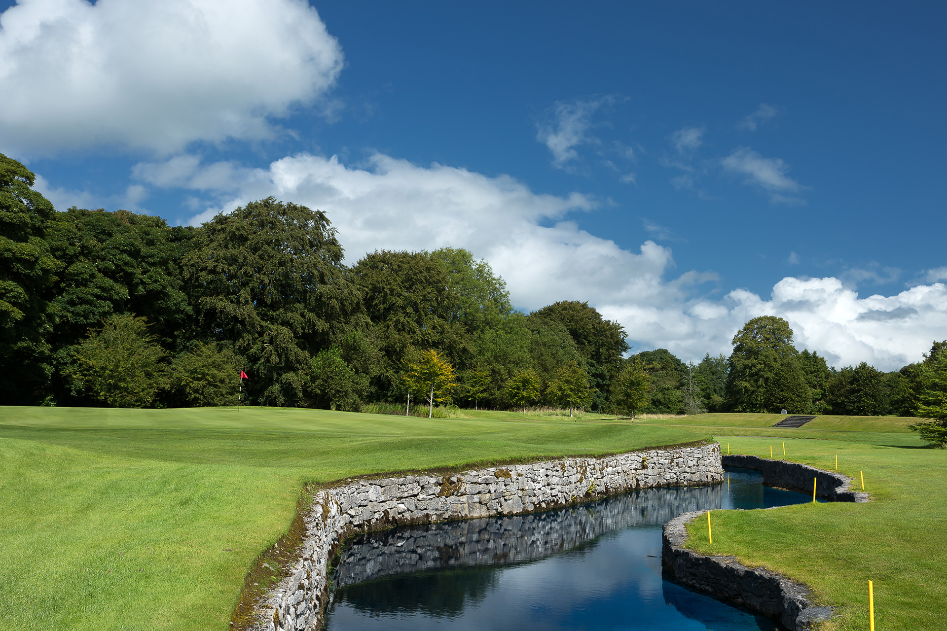 Golf course with a stone-lined water hazard curving alongside a well-maintained green with a red flag, surrounded by lush trees under a partly cloudy blue sky.