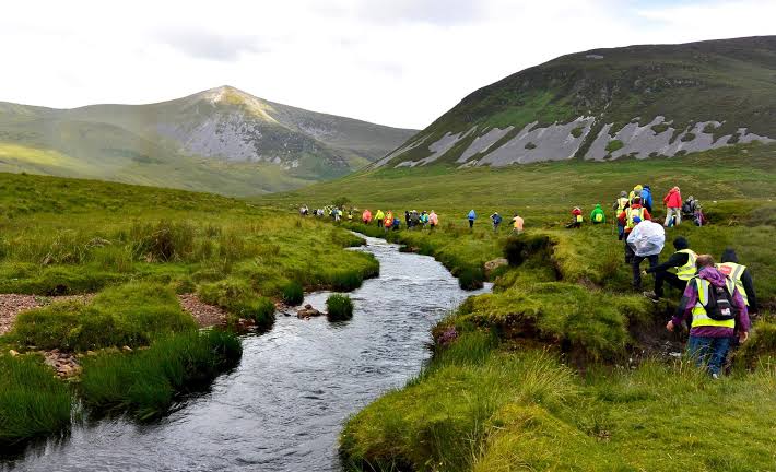 A group of hikers wearing colorful jackets walking along a grassy path next to a narrow river in a valley surrounded by green mountains under a cloudy sky.