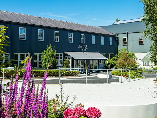 Exterior of Foxford Woollen Mills building with dark gray siding, multiple windows, an entrance canopy, and surrounded by greenery and flowering plants under a blue sky.