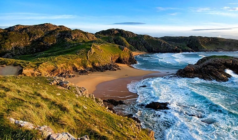Scenic coastal landscape showing green grassy hills, rocky cliffs, a sandy beach, and ocean waves crashing against the shore under a clear blue sky.