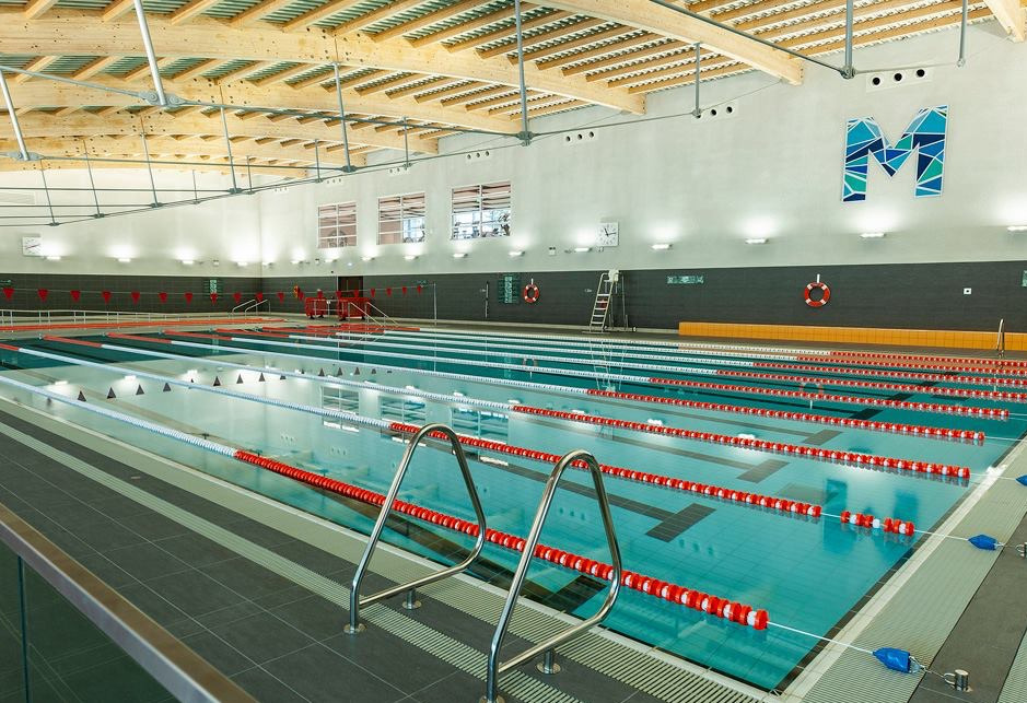 Indoor swimming pool with multiple lanes separated by red and white lane dividers, stainless steel pool ladder in foreground, wooden beam ceiling, large white walls with gray lower panels, clocks, life rings, and a blue geometric letter 'M' on the wall.