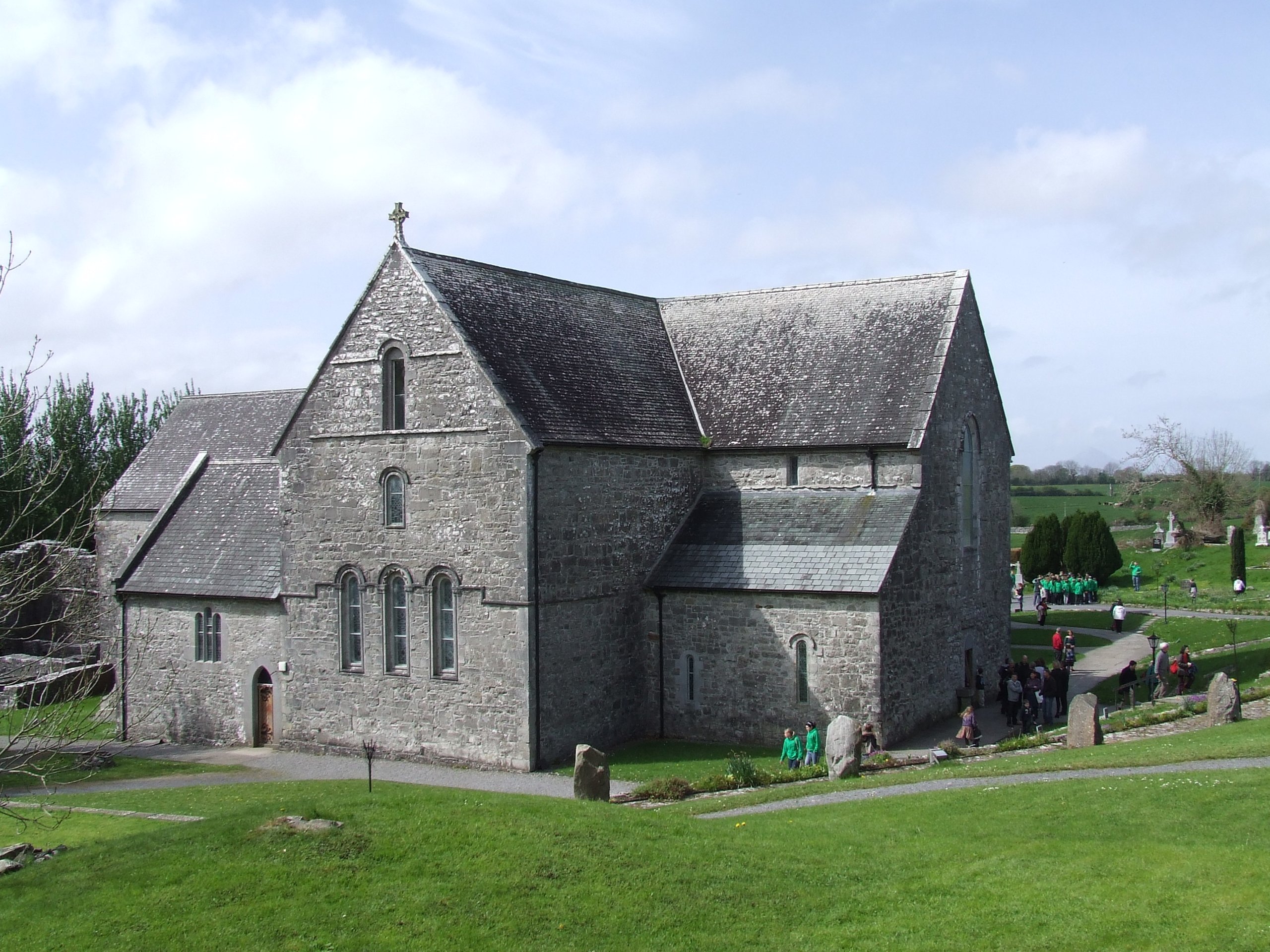 Gray stone church with a steep gabled roof and arched windows surrounded by green grass and pathways, with groups of people walking and standing nearby under a partly cloudy sky.