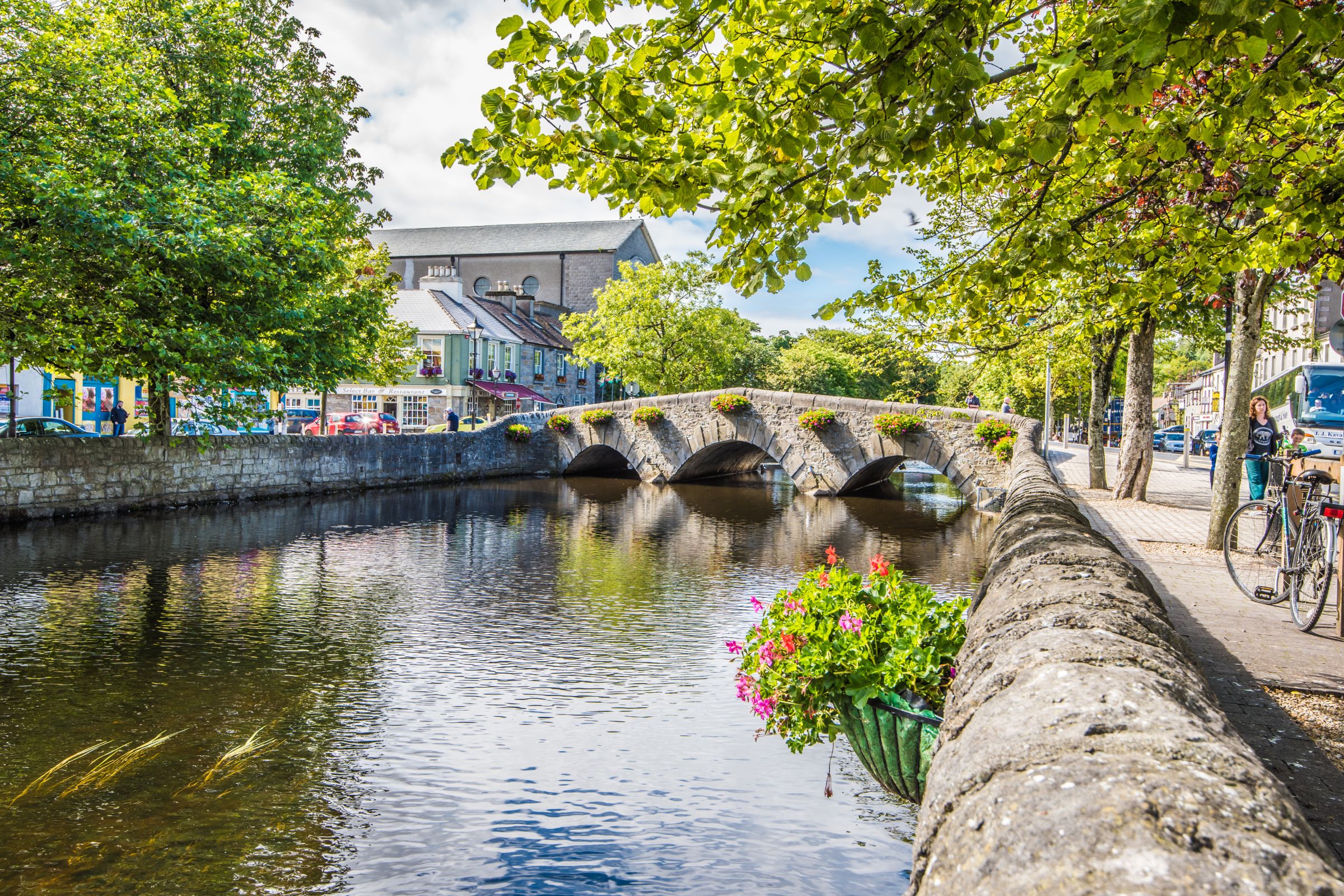 Stone bridge with three arches decorated with hanging flower baskets crossing a calm river, framed by leafy green trees on both sides, with a pedestrian walkway and parked bicycle on the right and colorful buildings in the background under a partly cloudy sky.