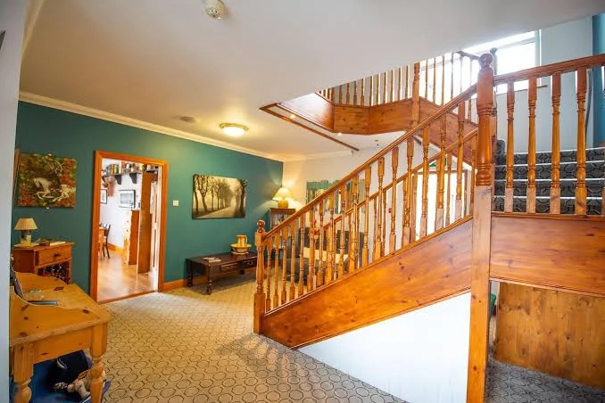 Bright foyer with wooden staircase featuring ornate spindles and carpeted steps, teal and white walls, patterned tile floor, wooden furniture, and framed landscape artwork.